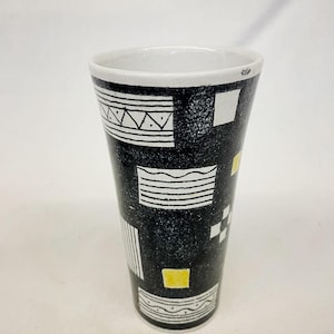 Op de afbeelding: Een hoge, zwarte keramische vaas met een witte binnenkant en een geometrisch patroon. Het ontwerp omvat witte en gele vierkanten, rechthoeken en driehoeken. De vaas heeft een gespikkelde textuur.