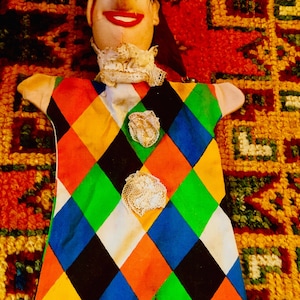 May include: A colorful puppet with a diamond pattern in red, orange, yellow, green, blue, and black. The puppet has a white face with a large, red, smiling mouth and a long, brown nose.