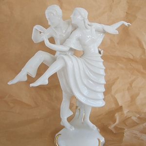 May include: A white porcelain figurine of a couple dancing. The woman is wearing a flowing dress and the man is holding her up. The figurine is on a white base with gold trim.