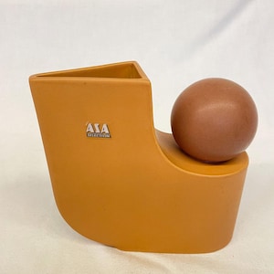 May include: A modern, orange-brown ceramic sculpture with a triangular opening and a rounded base. A brown sphere sits atop the base. The sculpture has the text "ASA SELECTION" on the front.