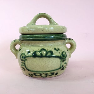 May include: A small, green and beige ceramic jar with a lid and a handle. The jar has a green floral design and a blank label.
