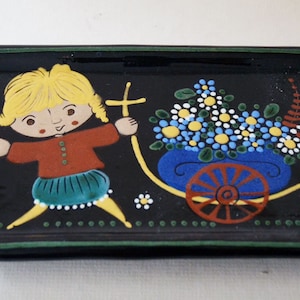 May include: A black ceramic rectangular dish with a green border. The dish features a hand-painted design of a girl pushing a cart full of flowers. The girl has blonde hair, a red shirt, a green skirt, and yellow shoes. The cart is blue with a red wheel.