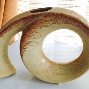May include: A ceramic vase with a unique, abstract design. The vase is a light beige color with brown accents and a large, circular opening.