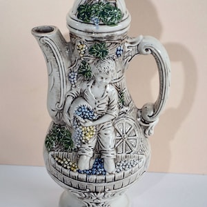 以下が含まれることがあります： ぶどうのつるからぶどうを摘んでいる少年の細かなデザインが施された白い陶器のピッチャー。ピッチャーは蓋と取っ手があり、緑の葉、茶色のつる、青と黄色のぶどうで飾られています。