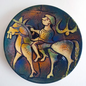 Puede incluir: Un plato redondo de cerámica con un diseño abstracto colorido de una persona montando a caballo. El plato tiene un fondo azul oscuro con detalles dorados y marrones.