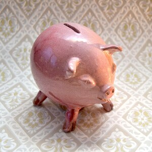 May include: A pink ceramic piggy bank with a slot on top for coins. The piggy bank has four short legs and a smiling face.