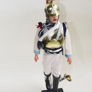 Puede incluir: Una figurita de soldado de juguete vintage que lleva un casco dorado con una pluma rosa, un uniforme blanco con detalles azules y rojos y botas negras. El soldado sostiene una espada y está de pie sobre una base negra con la palabra "CARABINIER" escrita en ella.