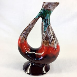 Puede incluir: Un jarrón de cerámica rojo, marrón y verde con un diseño abstracto único. El jarrón tiene una base ancha y un cuello estrecho, con un agujero en el lateral.