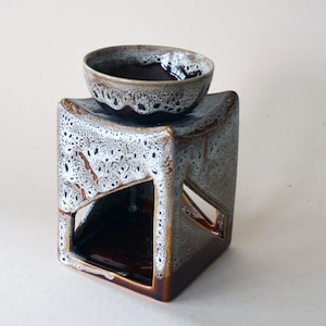 May include: A brown and white ceramic oil burner with a square base and a round bowl on top. The base has a cutout design and a speckled glaze.