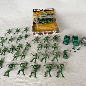 May include: A set of 32 green plastic toy soldiers in various poses, along with a tank, two jeeps, and a tank. The soldiers are arranged in rows on a white surface. The packaging for the toys is visible in the background, with the words "Combat Soldiers" and "32 Assorted Action Poses" printed on it.
