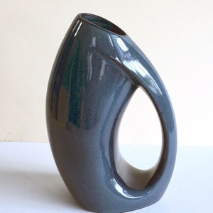 May include: A dark gray ceramic vase with a unique, abstract design. The vase has a large opening at the top and a cut-out handle.