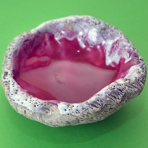 May include: A small, round ceramic bowl with a rough, textured exterior and a smooth, pink interior. The bowl has a white glaze around the rim.