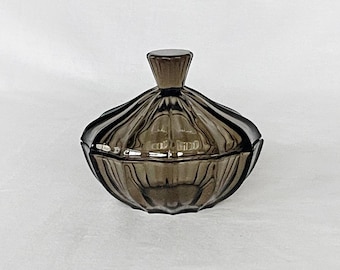 May include: A dark gray glass candy dish with a fluted design and a matching lid. The dish has a round shape and a slightly raised center.