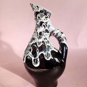 Puede incluir: Un jarrón de cerámica negro con un diseño de esmalte blanco. El esmalte tiene un efecto burbujeante y texturizado. El jarrón tiene una forma única y curvada con un asa.