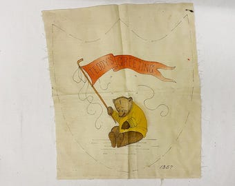 May include: A vintage cream-colored fabric with a brown teddy bear holding a red banner that says "Teddy's Greeting". The banner is attached to a brown stick. The fabric has a brown outline of a shield shape and the year "1957" is written in the bottom right corner.