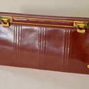 Könnte beinhalten: Eine braune Leder-Clutch mit einem goldenen Metallverschluss und Verzierungen. Die Clutch hat ein dezentes, strukturiertes Design und ein goldenes 'G'-Logo unten rechts.
