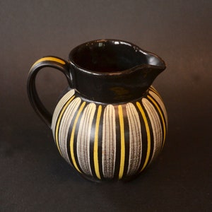 May include: A black ceramic pitcher with a yellow and white striped design. The pitcher has a rounded body and a handle.