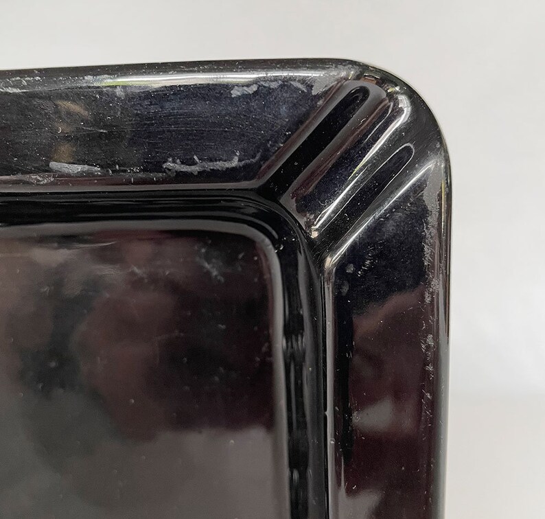 K&ouml;nnte beinhalten: Schwarzer Aschenbecher aus Glas mit quadratischer Form und abgeschr&auml;gter Kante. Der Aschenbecher hat eine glatte, gl&auml;nzende Oberfl&auml;che.