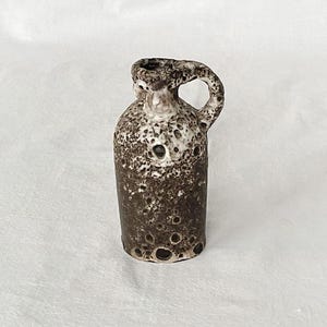 Puede incluir: Un pequeño jarrón de cerámica marrón y blanco con una superficie texturizada y un asa. El jarrón tiene un cuerpo redondo y un cuello estrecho.