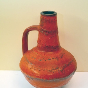 May include: A red ceramic vase with a textured surface and a handle. The vase has a wide base and a narrow neck.