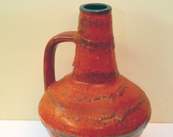 May include: A red ceramic vase with a textured surface and a handle. The vase has a wide base and a narrow neck.