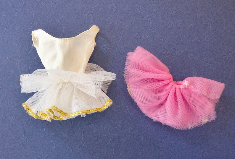 May include: Two tulle tutus for a doll. One is white with a gold trim and the other is pink.