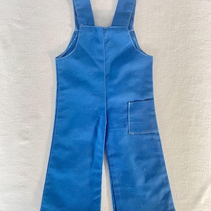 May include: Bright blue overalls with wide legs and adjustable straps. The garment features a single square pocket on the right leg. The fabric appears to be a sturdy cotton or blend, suitable for children's wear.
