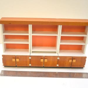 Op de afbeelding: Een houten rek met drie secties, elk met drie planken. De planken zijn wit met oranje accenten. De onderste sectie heeft drie lades met houten fronten en messing knoppen.