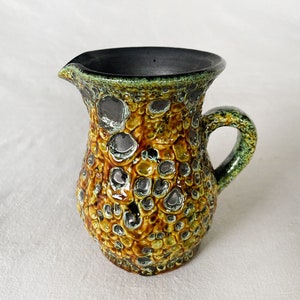 May include: A small, textured ceramic pitcher with a handle and spout. The pitcher features a mottled glaze in shades of brown, yellow, and green, with dark gray accents. The interior is black.