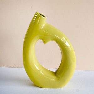 May include: A bright yellow ceramic vase with a unique heart-shaped opening. The vase is smooth and glossy, with a modern and abstract design.