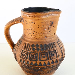 Puede incluir: Una jarra de cerámica marrón con un diseño geométrico negro. La jarra tiene una boca ancha y un asa.