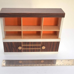 Op de afbeelding: Een bruin houten kastje met een witte rand en twee deuren. Het kastje heeft een plank met zes vakken en een onderste plank met twee deuren. De deuren hebben ronde knoppen.