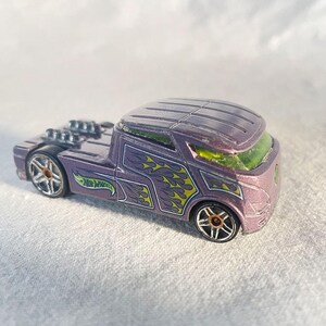 May include: Purple Hot Wheels toy car with a unique design. The car features flame graphics in yellow and green, along with the Hot Wheels logo. The car has a distinctive cab and exposed engine components.