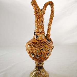 May include: A brown and yellow ceramic vase with a textured surface and a unique, organic shape. The vase has a wide base and a narrow neck with a handle.