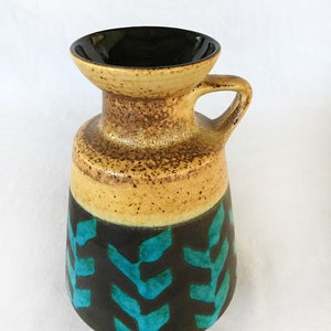 Pode incluir: Um vaso de cerâmica com uma alça, apresentando um interior preto e um exterior bege salpicado. A base é preta com padrões turquesa em forma de folha. O vaso tem um design artístico único.