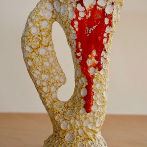 May include: A ceramic pitcher with a textured surface and a red glaze. The pitcher has a handle and a signature that reads "Rom."