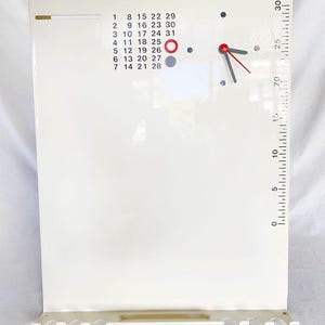 Pode incluir: Um calendário de parede branco com um relógio e uma escala tipo régua na lateral. O calendário tem números de 1 a 31, e o relógio tem ponteiros vermelhos. Uma pequena prateleira na parte inferior.