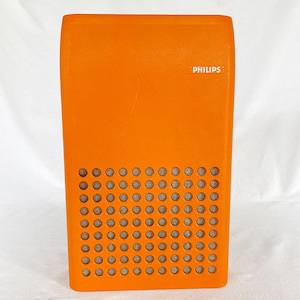 以下が含まれることがあります： オレンジ色のヴィンテージPhilipsラジオ。スピーカーグリルには小さな丸い穴がたくさんあります。ブランド名「PHILIPS」が白で印刷されています。ラジオは角が丸い長方形です。