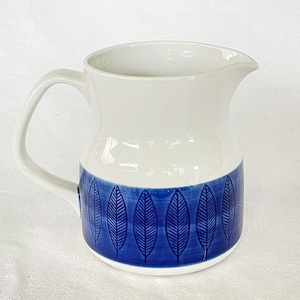 May include: White ceramic pitcher with a blue leaf pattern around the base. The pitcher has a curved handle and a spout for easy pouring. A decorative piece for serving beverages.