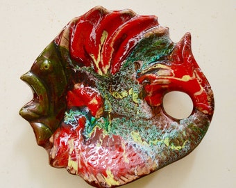 May include: A red, green, and white ceramic fish-shaped dish with a hole in the side. The dish has a mottled glaze and a textured surface.