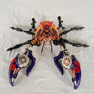 May include: A crab-shaped toy with a metallic orange and gold body, black legs, and purple and red claws. The toy has intricate mechanical details and is posed on a white surface.