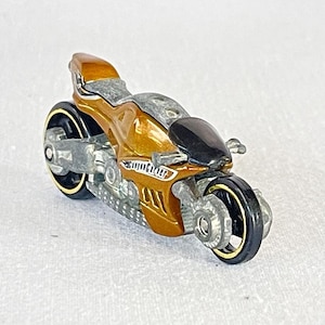 May include: A gold and black toy motorcycle with a silver frame and black wheels. The motorcycle has a detailed design with a black seat and a silver exhaust pipe.