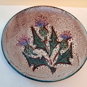 May include: A ceramic plate with a brown and white speckled glaze. The plate features a design of three stylized purple and pink thistles with green leaves.