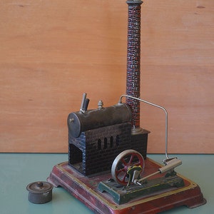 Puede incluir: Una máquina de vapor de juguete vintage con una chimenea de ladrillo rojo y negro, una caldera negra y una base verde. La máquina tiene un volante de inercia de metal plateado y una biela de metal plateado. La máquina está sobre una superficie verde.