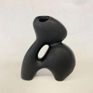 May include: A matte black ceramic vase with an abstract, organic form. The vase features a rounded base, a central opening, and a top opening. The design is modern and sculptural, suitable for home decor.