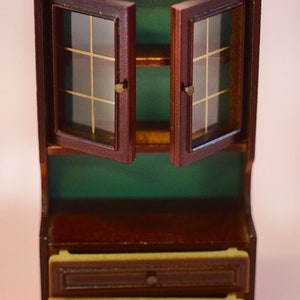 Op de afbeelding: Een miniatuur houten kast met twee glazen deuren, een plank en twee laden. De kast is bruin geverfd met een groene binnenkant.