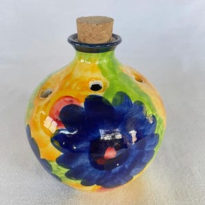 Puede incluir: Una botella de aceite de cerámica pintada a mano con un tapón de corcho. La botella presenta un diseño floral vibrante con una gran flor azul y acentos de amarillo, verde y rojo. La botella tiene varios agujeros pequeños.