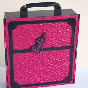 May include: A pink and black plastic toy suitcase with a handle and a Barbie logo. The suitcase has a diamond pattern on the front.