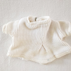 Puede incluir: Una camiseta blanca de punto de gofre con mangas cortas. La camiseta tiene un cuello alto y redondo y una superficie texturizada. La tela parece suave y ligera, adecuada para capas o clima cálido. La prenda está tendida.