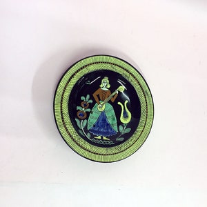 May include: A decorative plate with a green and black border. The center of the plate features a woman in a blue and green dress playing a musical instrument. The background is dark blue with floral designs.